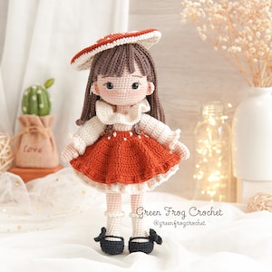Puede incluir: Una mu&ntilde;eca de crochet con un vestido rojo y blanco, una peluca marr&oacute;n y un sombrero de hongo rojo. La mu&ntilde;eca est&aacute; de pie sobre una superficie blanca con un peque&ntilde;o cactus y un jarr&oacute;n en el fondo. Green Frog Crochet @greenfrogcrochet