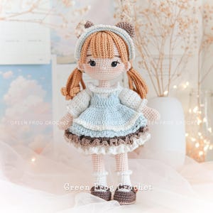 Peut inclure: Une poup&eacute;e en crochet avec des cheveux bruns, une robe bleue et des chaussures brunes. La poup&eacute;e porte un bandeau blanc avec des oreilles d'ours brunes. La poup&eacute;e est debout sur une surface blanche avec un fond blanc.