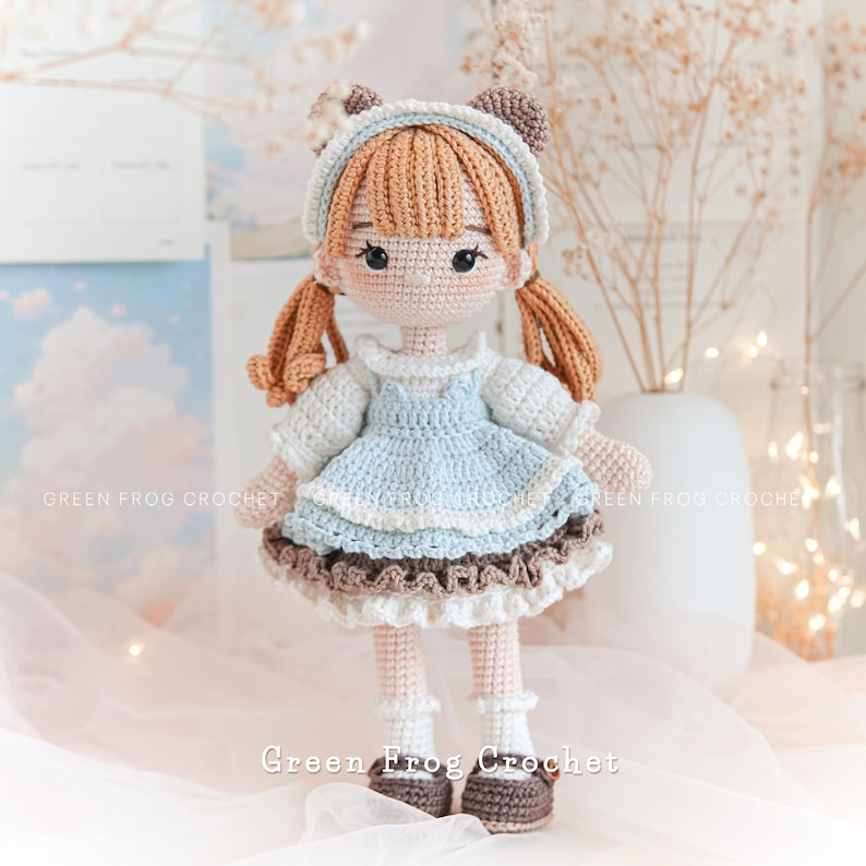 Peut inclure: Une poup&eacute;e en crochet avec des cheveux bruns, une robe bleue et des chaussures brunes. La poup&eacute;e porte un bandeau blanc avec des oreilles d'ours brunes. La poup&eacute;e est debout sur une surface blanche avec un fond blanc.