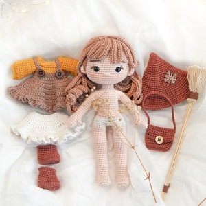 May include: Crocheted doll with brown hair and a pink dress, along with a set of crocheted clothing accessories including a white skirt, a brown dress, a brown purse, brown boots, and a red hooded cloak. The doll is on a white fabric background.