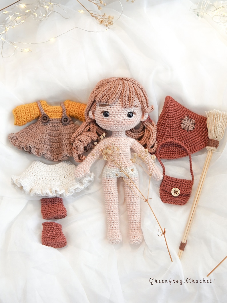 May include: Crocheted doll with brown hair and a pink dress, along with a set of crocheted clothing accessories including a white skirt, a brown dress, a brown purse, brown boots, and a red hooded cloak. The doll is on a white fabric background.