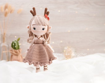 May include: A crocheted doll with brown hair, wearing a brown dress and antlers. The doll has a red flower on its head and is sitting on a white surface.