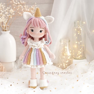 Puede incluir: Una mu&ntilde;eca de unicornio de crochet con un vestido rosa y blanco con rayas de arco&iacute;ris. La mu&ntilde;eca tiene una melena rosa y blanca y un cuerno rosa, y lleva zapatos blancos.