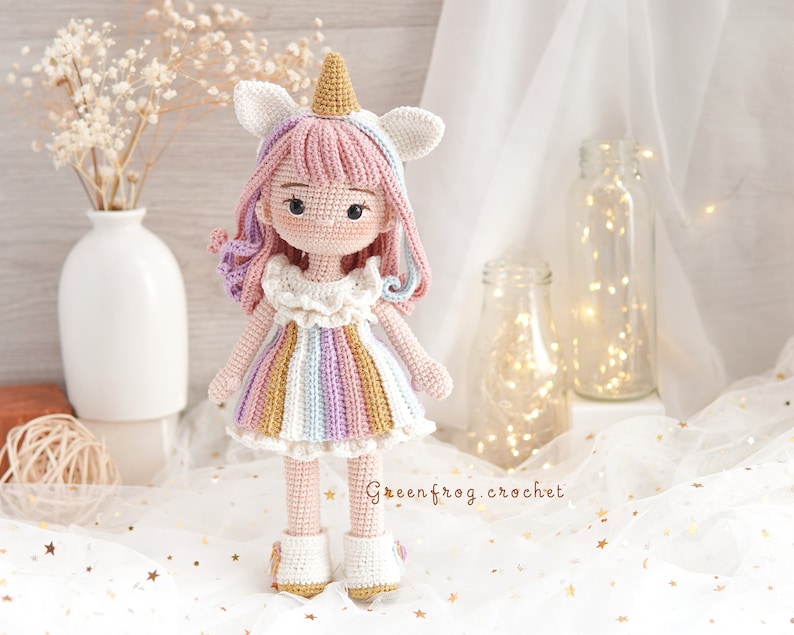 Puede incluir: Una mu&ntilde;eca de unicornio de crochet con un vestido rosa y blanco con rayas de arco&iacute;ris. La mu&ntilde;eca tiene una melena rosa y blanca y un cuerno rosa, y lleva zapatos blancos.