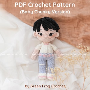 Puede incluir: Muñeca de ganchillo con cabello negro, que lleva una camisa blanca, pantalones azul claro y zapatos blancos con ribete rosa. La muñeca está sobre un fondo de tela blanca y beige. El texto de la imagen dice "PDF Crochet Pattern (Baby Chunky Version)" y "by Green Frog Crochet."