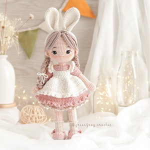 Puede incluir: Una mu&ntilde;eca de crochet que lleva un vestido blanco con un ribete de volantes rosa y una diadema de orejas de conejo. La mu&ntilde;eca tiene el pelo casta&ntilde;o y los ojos marrones. La mu&ntilde;eca est&aacute; de pie sobre una superficie blanca.