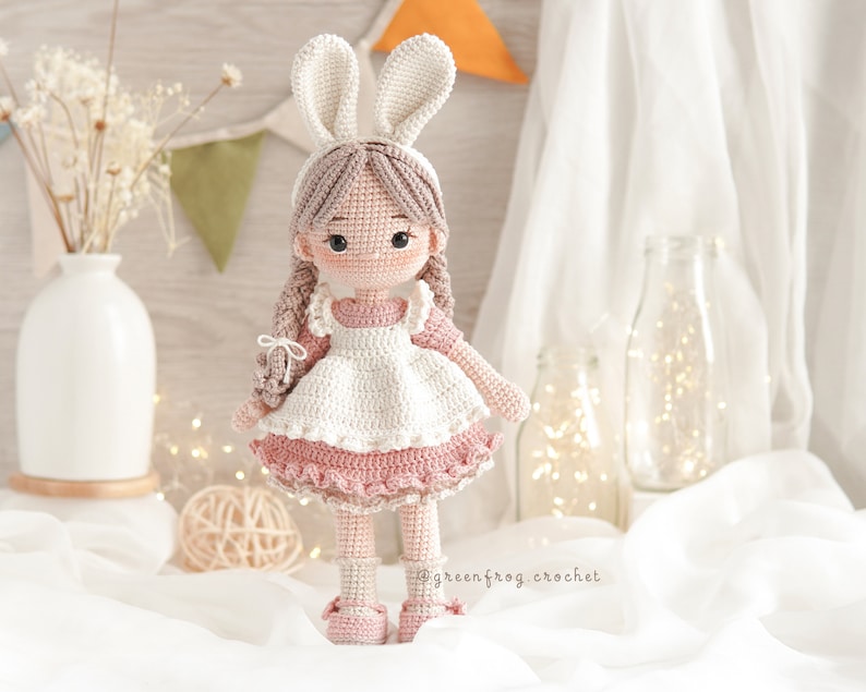 Puede incluir: Una mu&ntilde;eca de crochet que lleva un vestido blanco con un ribete de volantes rosa y una diadema de orejas de conejo. La mu&ntilde;eca tiene el pelo casta&ntilde;o y los ojos marrones. La mu&ntilde;eca est&aacute; de pie sobre una superficie blanca.