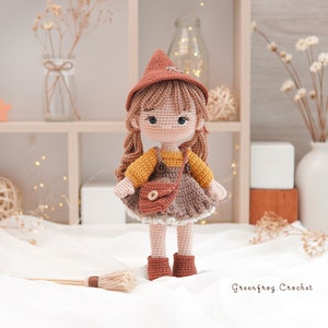 May include: A crocheted doll wearing a brown witch hat, yellow and brown dress, and brown boots. The doll is holding a brown purse and is standing on a white surface with a broom in front of it. The text "Greenfrog Crochet" is visible in the bottom right corner of the image.