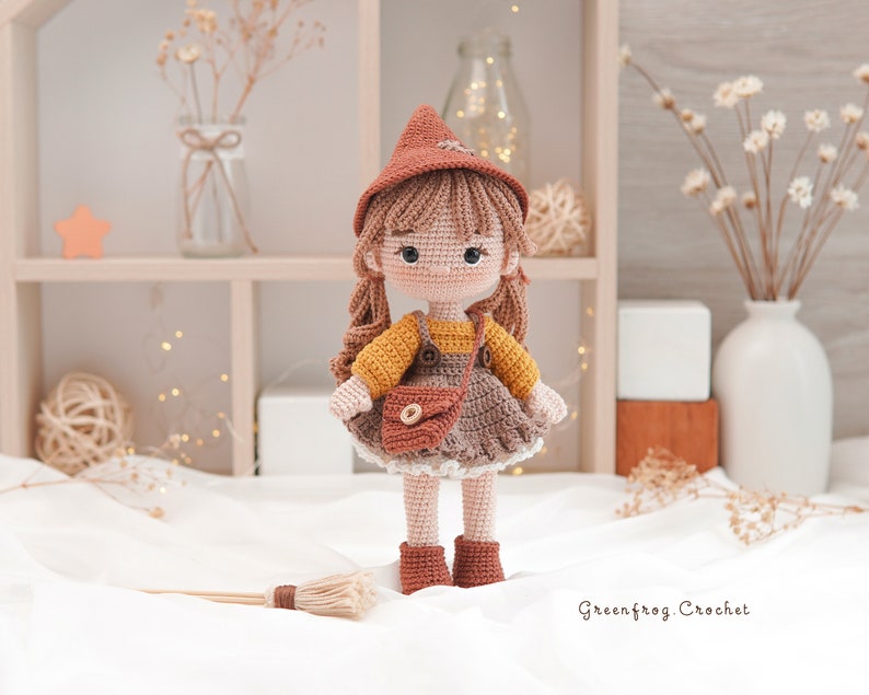 May include: A crocheted doll wearing a brown witch hat, yellow and brown dress, and brown boots. The doll is holding a brown purse and is standing on a white surface with a broom in front of it. The text "Greenfrog Crochet" is visible in the bottom right corner of the image.