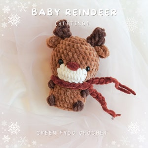 May include: A brown crocheted reindeer toy with a red scarf, sitting on a white background. The toy has a white face with black eyes and a red nose. The text "BABY REINDEER (SITTING)" is above the toy, and the text "GREEN FROG CROCHET" is below the toy.