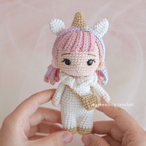 Puede incluir: Una mu&ntilde;eca de unicornio de crochet con pelo rosa, un vestido blanco y un bolso dorado. La mu&ntilde;eca lleva un cuerno y orejas de unicornio.