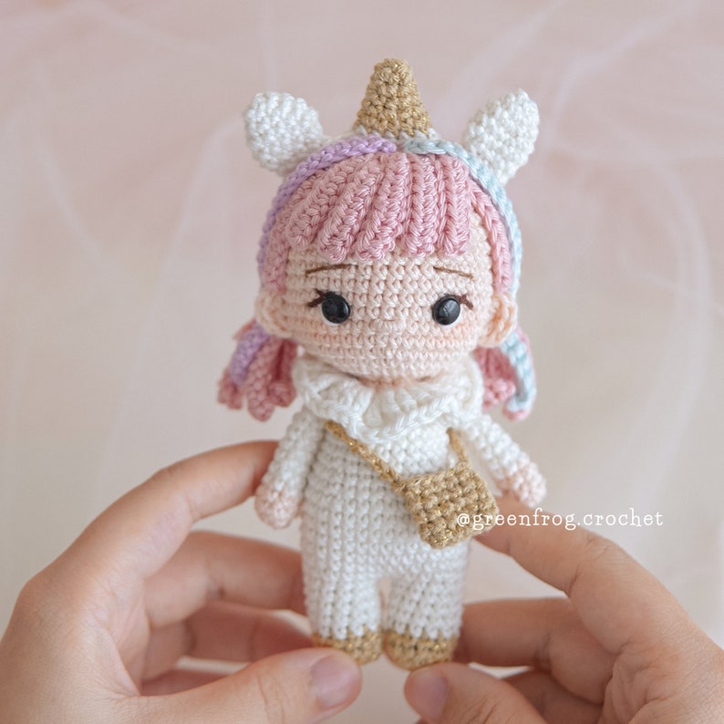 Puede incluir: Una mu&ntilde;eca de unicornio de crochet con pelo rosa, un vestido blanco y un bolso dorado. La mu&ntilde;eca lleva un cuerno y orejas de unicornio.