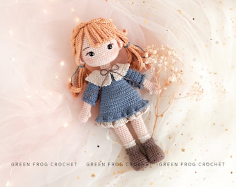 Könnte beinhalten: Eine gehäkelte Puppe mit braunen Haaren, einem blauen Kleid und braunen Schuhen. Die Puppe sitzt auf einem weißen Hintergrund mit goldenem Glitzer.