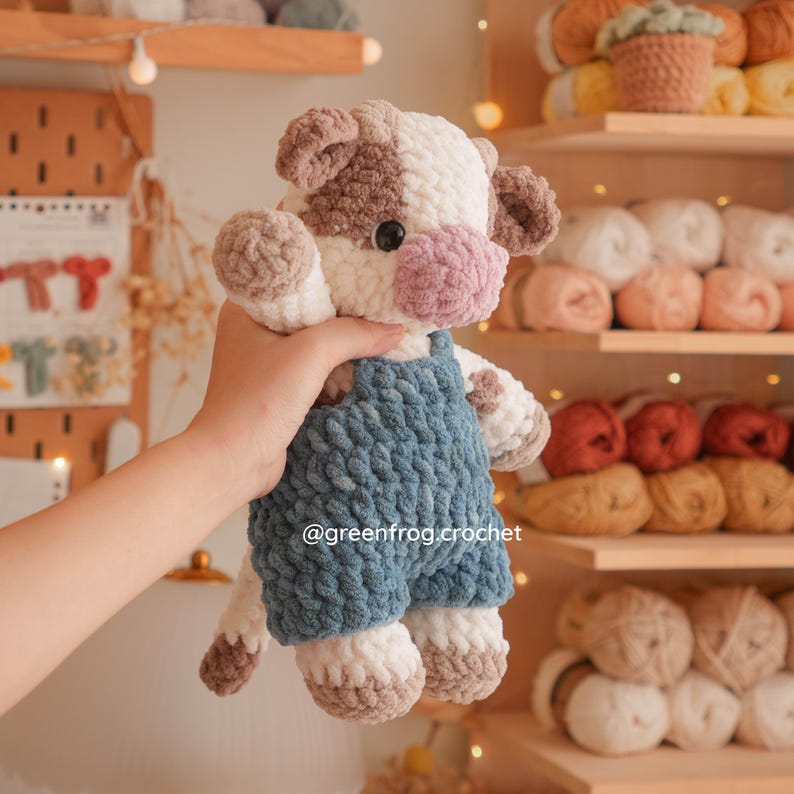 May include: A plush cow toy made of soft white and brown yarn, wearing blue crocheted overalls. The cow has a pink snout and black eyes. The text @greenfrog.crochet is visible on the blue overalls. The toy is crafted with a thick, textured stitch.