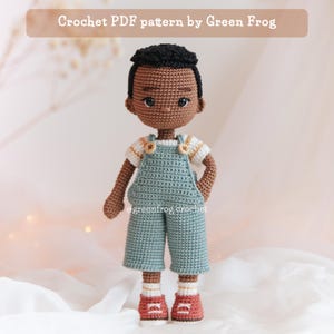 Puede incluir: Muñeco de ganchillo de un niño pequeño con una camisa a rayas blancas y amarillas, un peto azul y zapatillas rojas. El muñeco tiene pelo negro y piel morena. El texto "Crochet PDF pattern by Green Frog" está en la parte superior.