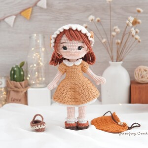 May include: A crocheted doll with brown hair, a white dress, and a white hat. The doll is holding a small brown basket. The doll is standing on a white surface with a brown bag and a vase of dried flowers in the background.