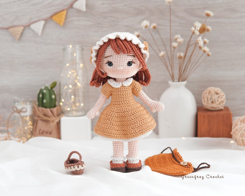 May include: A crocheted doll with brown hair, a white dress, and a white hat. The doll is holding a small brown basket. The doll is standing on a white surface with a brown bag and a vase of dried flowers in the background.