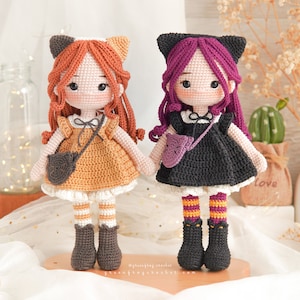 Puede incluir: Dos mu&ntilde;ecas de crochet con orejas de gato. Una mu&ntilde;eca lleva un vestido marr&oacute;n con una falda blanca y medias a rayas blancas y naranjas. La otra mu&ntilde;eca lleva un vestido negro con una falda blanca y medias a rayas naranjas y moradas. Ambas mu&ntilde;ecas llevan zapatos negros.