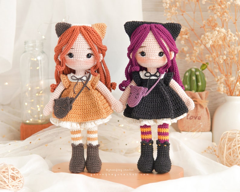 Puede incluir: Dos mu&ntilde;ecas de crochet con orejas de gato. Una mu&ntilde;eca lleva un vestido marr&oacute;n con una falda blanca y medias a rayas blancas y naranjas. La otra mu&ntilde;eca lleva un vestido negro con una falda blanca y medias a rayas naranjas y moradas. Ambas mu&ntilde;ecas llevan zapatos negros.