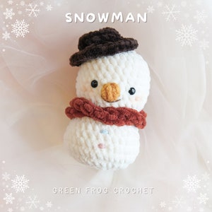 May include: A white crocheted snowman wearing a brown hat and a red scarf. The snowman has black eyes, an orange nose, and a smiling mouth. The background is a pink and white tulle fabric with white snowflakes.