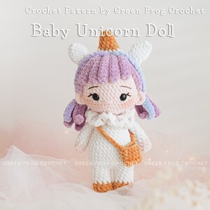 Puede incluir: Una mu&ntilde;eca de beb&eacute; unicornio de crochet con una melena de color p&uacute;rpura y blanco, un cuerno blanco con una punta naranja y un vestido blanco con un bolso marr&oacute;n. La mu&ntilde;eca est&aacute; de pie sobre una superficie rosa.