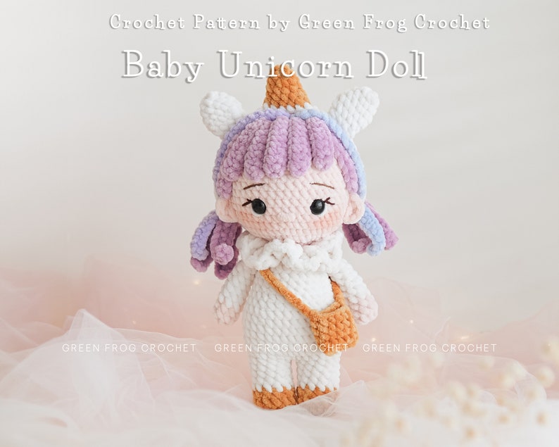 Puede incluir: Una mu&ntilde;eca de beb&eacute; unicornio de crochet con una melena de color p&uacute;rpura y blanco, un cuerno blanco con una punta naranja y un vestido blanco con un bolso marr&oacute;n. La mu&ntilde;eca est&aacute; de pie sobre una superficie rosa.
