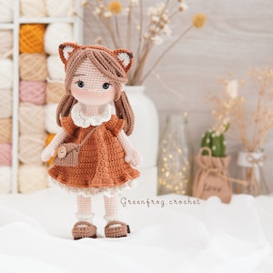 Puede incluir: Una mu&ntilde;eca de crochet con cabello casta&ntilde;o, vestida con un vestido marr&oacute;n con cuello blanco y un bolso marr&oacute;n. La mu&ntilde;eca lleva zapatos marrones y tiene una cara de zorro con orejas marrones.