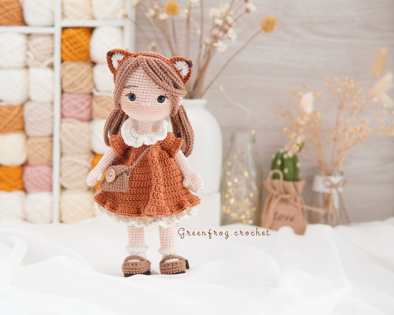 Puede incluir: Una mu&ntilde;eca de crochet con cabello casta&ntilde;o, vestida con un vestido marr&oacute;n con cuello blanco y un bolso marr&oacute;n. La mu&ntilde;eca lleva zapatos marrones y tiene una cara de zorro con orejas marrones.