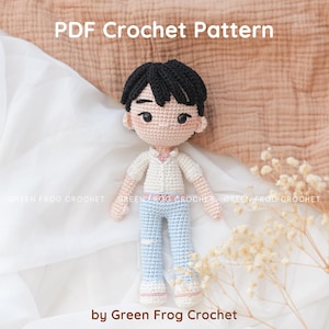 Może przedstawiać: Ręcznie robiona szydełkowa lalka z czarnymi włosami, ubrana w białą koszulę, jasnoniebieskie spodnie i trampki. Lalka jest umieszczona na miękkim, neutralnym tle. Tekst na zdjęciu to "PDF Crochet Pattern" i "by Green Frog Crochet".