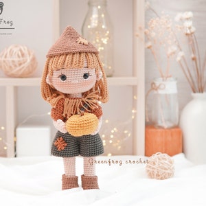 May include: Crocheted doll wearing a brown hat and holding a small orange pumpkin. The doll is wearing a brown and green outfit and has brown hair. The doll is standing on a white surface.