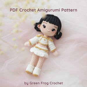 May include: A crocheted amigurumi doll with black hair, wearing a white and gold outfit. The doll has a detailed face with blush and earrings. The text "PDF Crochet Amigurumi Pattern" and "by Green Frog Crochet" are visible.