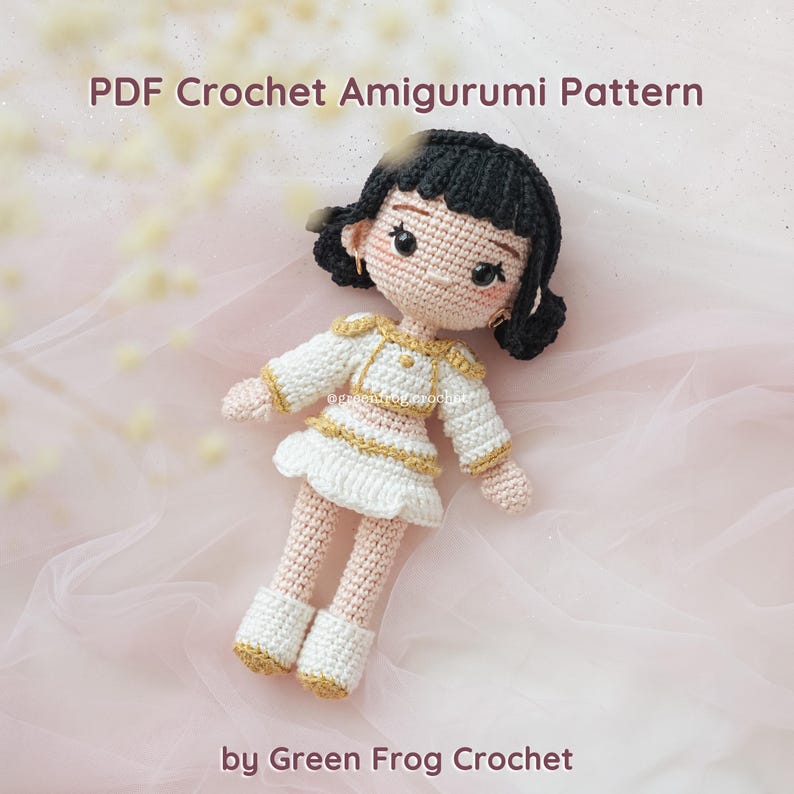 May include: A crocheted amigurumi doll with black hair, wearing a white and gold outfit. The doll has a detailed face with blush and earrings. The text "PDF Crochet Amigurumi Pattern" and "by Green Frog Crochet" are visible.