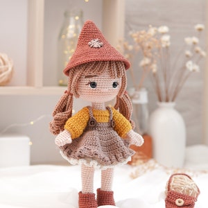 May include: A crocheted doll wearing a brown hat with a flower, a yellow and brown dress, and brown boots. The doll has brown hair in pigtails and brown eyes. The doll is standing on a white surface with a small crocheted bag next to it.