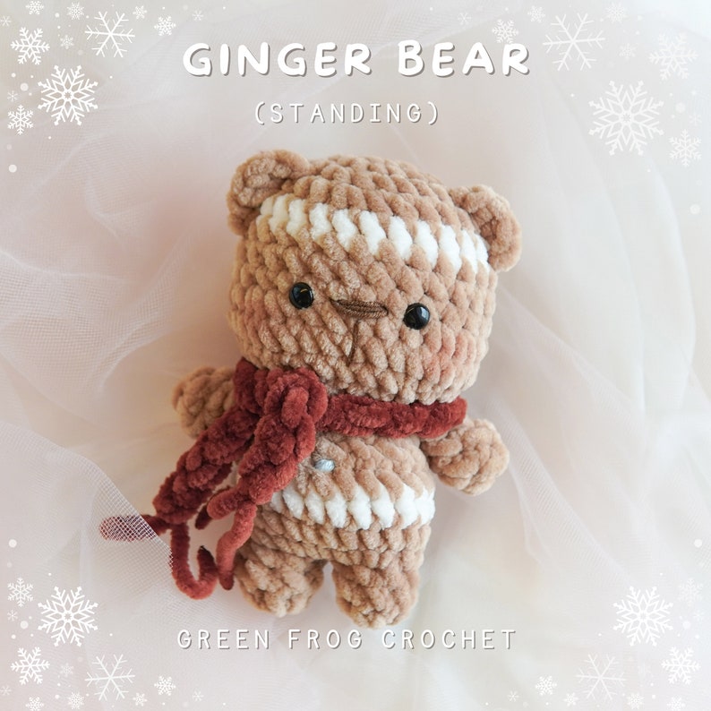 May include: A crocheted gingerbread bear toy standing on a white background. The bear is brown with white stripes and has a red scarf. The text "Ginger Bear (Standing)" is above the bear and the text "Green Frog Crochet" is below the bear.