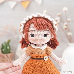 May include: A crocheted doll with brown hair, wearing a white and brown dress with a brown belt and a button. The doll has brown eyes and a white crocheted collar.