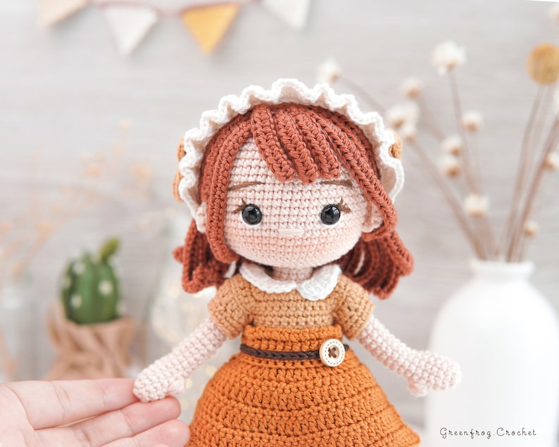 May include: A crocheted doll with brown hair, wearing a white and brown dress with a brown belt and a button. The doll has brown eyes and a white crocheted collar.