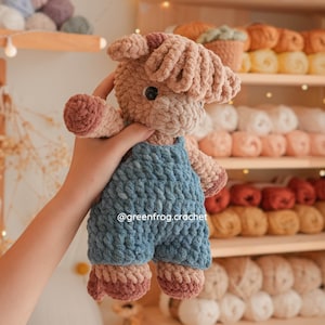 May include: A plush crochet toy of a highland cow with shaggy light brown yarn, dark brown hooves, and a blue yarn overall outfit. The toy features black plastic eyes and the text @greenfrog.crochet is visible on the blue overalls.