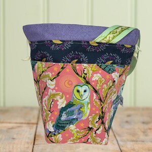 May include: A patterned tote bag with an owl motif. The bag has a pink base with a design of branches, leaves, and stars. The owl is shown with blue, green, and yellow plumage. The top of the bag has a purple and navy blue trim.