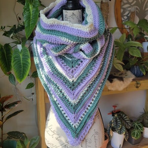 May include: A crocheted triangular scarf in shades of green, purple, and white. The scarf is draped over a mannequin's neck and shoulders.