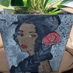 May include: Square black and gray planter pot with a painted portrait of a woman. The woman has dark hair, red lips, and a red rose. The pot is filled with soil and a green plant.