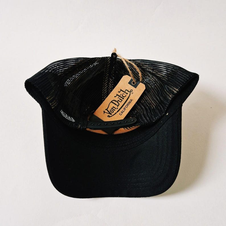 Puede incluir: Gorra de camionero negra con parte trasera de malla y visera curva. Una etiqueta marr&oacute;n con el texto "Von Dutch California" est&aacute; sujeta con hilo. La gorra se ve desde abajo.