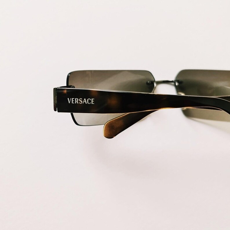 May include: Brown tortoiseshell sunglasses with a square frame and gray lenses. The sunglasses have the word "VERSACE" printed on the side.