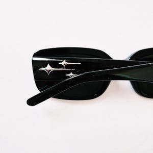 K&ouml;nnte beinhalten: Schwarze Sonnenbrille mit rechteckigem Rahmen und silbernen Stern-Akzenten. Die Sonnenbrille hat einen schwarzen Rahmen und Gl&auml;ser.