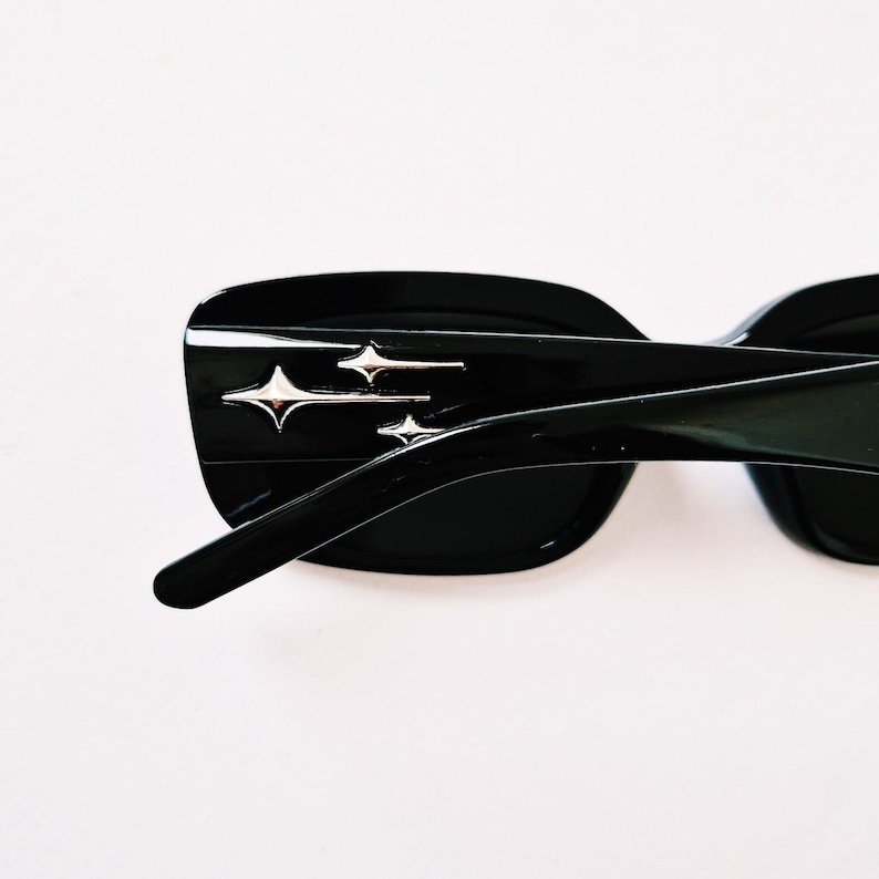 K&ouml;nnte beinhalten: Schwarze Sonnenbrille mit rechteckigem Rahmen und silbernen Stern-Akzenten. Die Sonnenbrille hat einen schwarzen Rahmen und Gl&auml;ser.