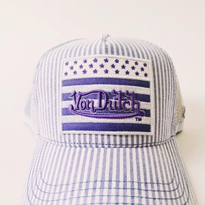 May include: A blue and white striped trucker hat with a purple embroidered patch. The patch features the text "Von Dutch" in a stylized font, along with a design of stars and stripes. The hat has a mesh back.