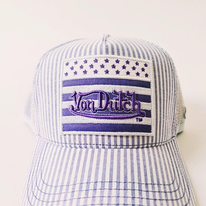 May include: A blue and white striped trucker hat with a purple embroidered patch. The patch features the text "Von Dutch" in a stylized font, along with a design of stars and stripes. The hat has a mesh back.