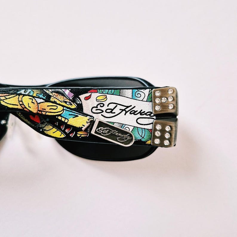 Może przedstawiać: Czarne okulary przeciwsłoneczne z kolorowym wzorem Ed Hardy przedstawiającym węża i akcenty w postaci kości do gry. Okulary mają czarną oprawkę i szkła.