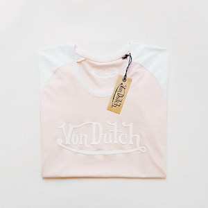 May include: A folded pink and white raglan t-shirt with the text "Von Dutch" in white script. A brown tag with the "Von Dutch" logo hangs from the collar. The shirt is folded neatly on a white surface.