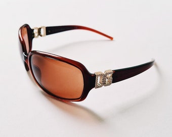 Puede incluir: Gafas de sol marrones con un logotipo DG dorado en el lateral. Las lentes son marrones y la montura es de plástico.