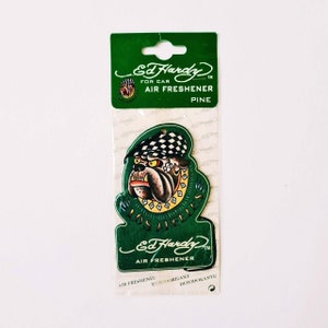 Op de afbeelding: Groene en zwarte Ed Hardy autoluchtverfrisser met een bulldog die een geruite hoed en een gouden ketting draagt. De luchtverfrisser is gevormd als een bulldoghoofd en heeft de woorden "Los Angeles" erop geschreven. De luchtverfrisser is gemarkeerd met "Pijnboom".